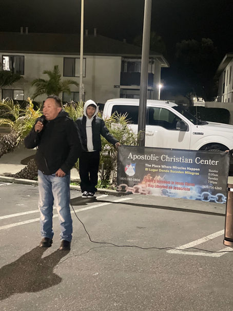 Man speaking, another standing nearby, "Assemblies of God Church" banner