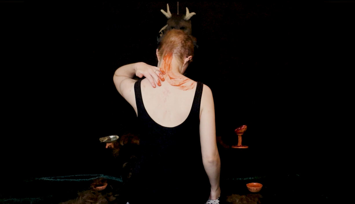 Yveline Burton, a white femme person with red hair and a black tank top on, sits with her back to the camera. Her hair is shaved unevenly but completely. She is using her left hand to smear red-orange liquid clay down from the nape of her neck and over her left shoulder blade like wings. a trail of clay shows she had sone the same on the other side of her body already.  She is surrounded by her shaved hair spilling off her lap and fallen to either side of her, and by small ritual objects, terracotta bowls, a shiny ormer shell with incense in it, and a halved pomegranate dripping juice down a terracotta stand. Behind her head, suspended in the top middle of the frame, we see the very top of an animalistic, brown furry mask with sheep-like horns peeking out. 
