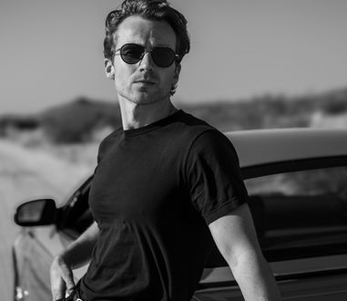 Black and white photo of a man wearing glasses standing and leaning against a car in the desert.