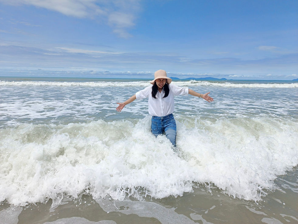 Smiling woman in a sunhat and white shirt stands in foamy surf with arms out on a bright blue beach.