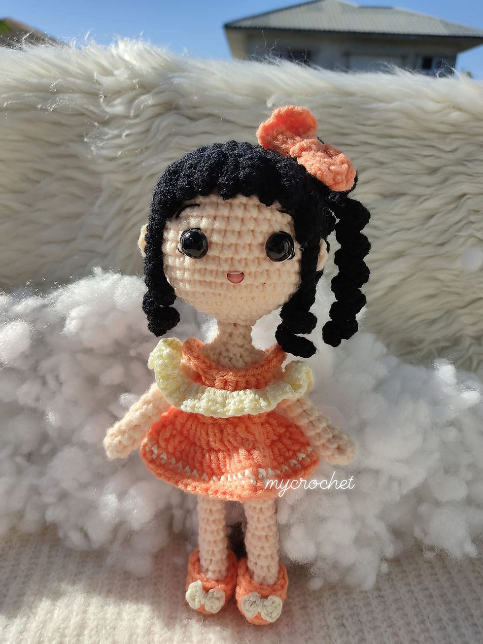 Crocheted girl doll with black braids, orange bow and peach dress stands on fluffy white blanket under blue sky; mycrochet watermark.