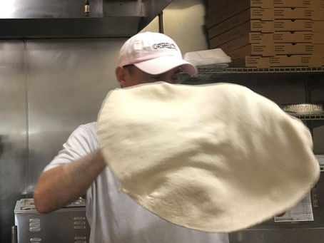 Pizza chef tossing dough in kitchen, wearing a pink cap and white shirt. A professional kitchen, with a stack of pizza boxes in the background, hood and some equipment
