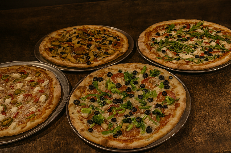 Four freshly baked gourmet pizzas on silver trays, displayed on a rustic wooden table. Each pizza is topped with fresh ingredients including jalapeños, black olives, broccoli, arugula, and cheese