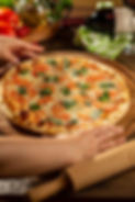 Freshly baked Margherita pizza topped with sliced tomatoes, mozzarella, and basil leaves on a wooden serving board, with hands presenting it near a Castello’s wine bottle, olive oil, and fresh ingredients