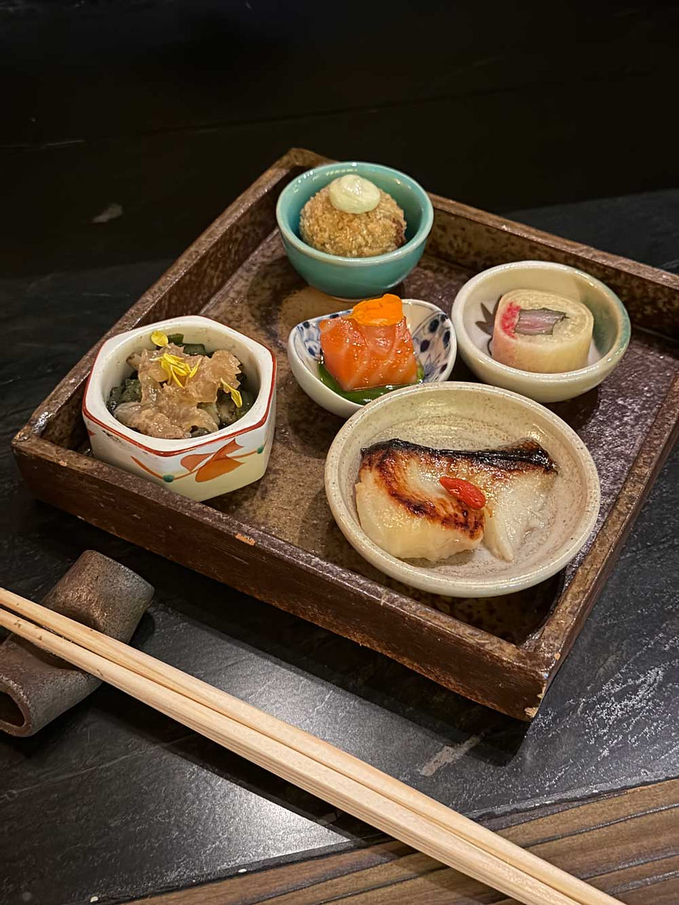 A wooden tray with assorted Japanese dishes in small bowls. Pair of wooden chopsticks are beside it.