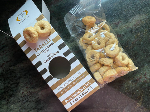Open box and plastic bag of cheese and pepper taralli on dark surface. Text: "Taralli, Cheese & Pepper, Cacio e Pepe, 150g". Light mood.