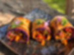 Three stuffed eggplant rolls with colorful veggies, garnished with purple onion flowers and green fresh cilantro leaves, served on a textured dark plate.