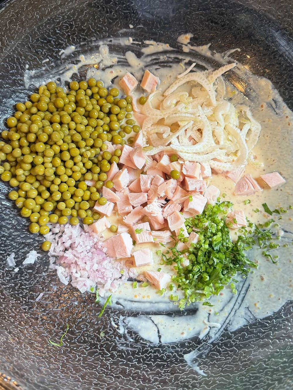 Canned green peas, diced smoked turkey breast, shallots, fennel bulb, and cilantro stems chopped in a bowl with a creamy dressing, creating a fresh, colorful salad mix.