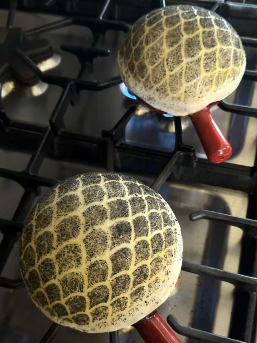 Two red terracotta pots are on the stove over medium heat. Their content covered with inflated dough decorated with black powder scales.