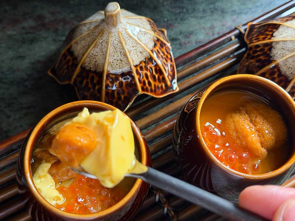 A spoon holds creamy egg custard with uni over a pot of chawanmushi with ikura. Brown eggplant-like lids and wooden mat in the background.
