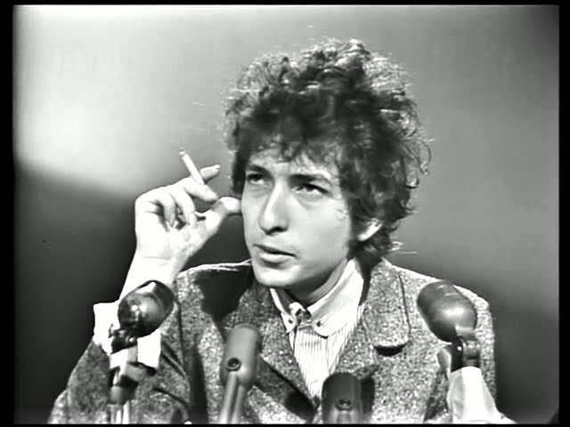 Curly haired man in black and white with cigarette sits at desk in front of microphones