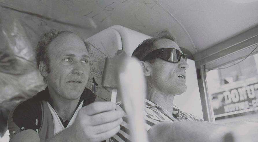 Man holding rolled cigarette next to man in dark sunglasses driving bus