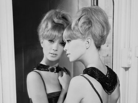 Black and white photo of blonde woman with updo looking into mirror