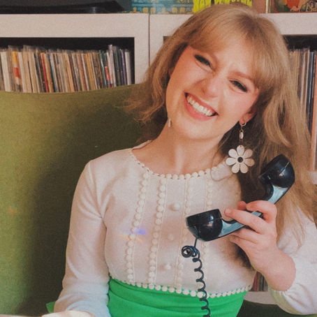 Blonde woman in white and green dress seated in green egg chair smiling with vinyl record and black telephone