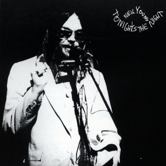 High contrast black and white image of long-haired man at microphone wearing dark sunglasses and seersucker jacket