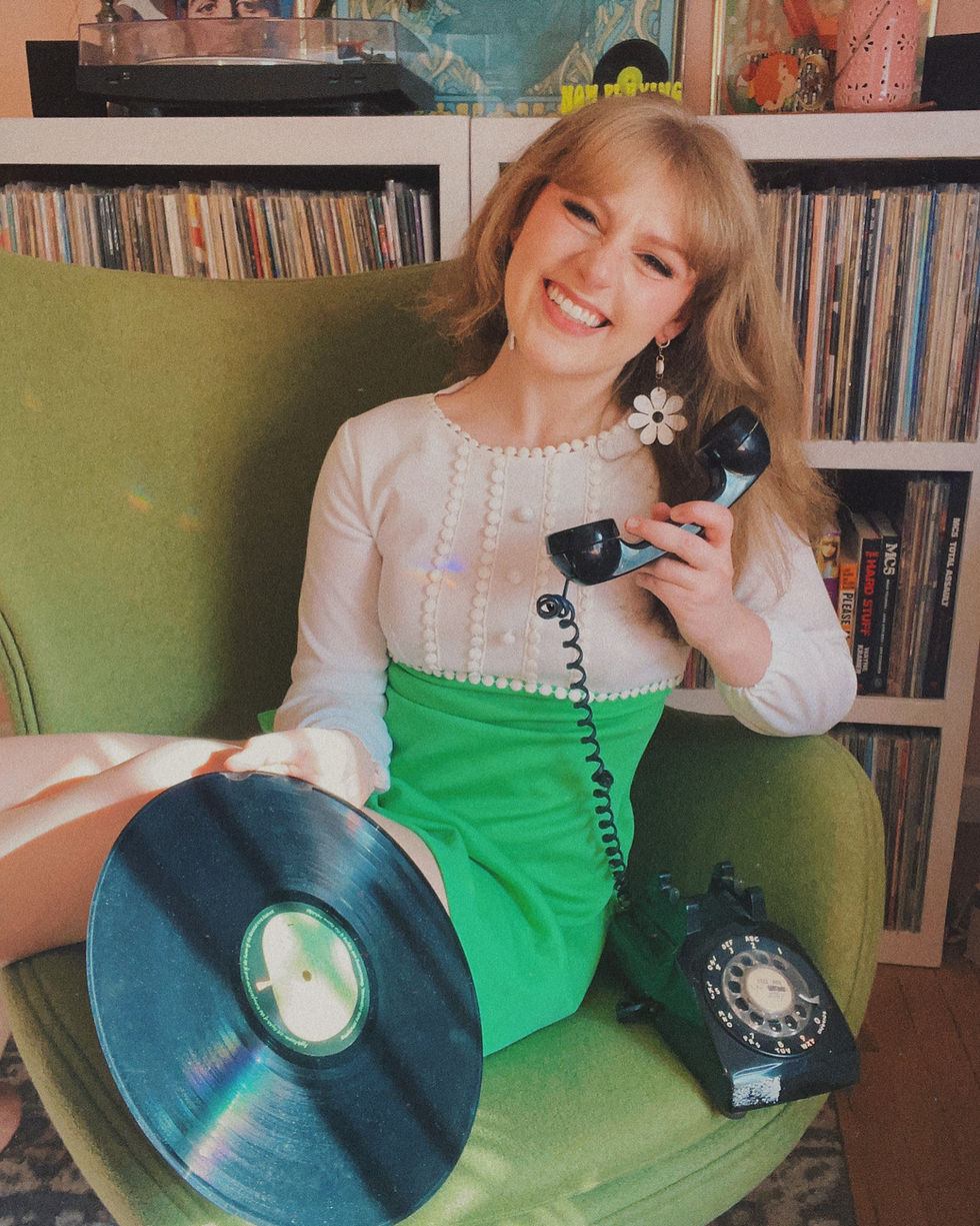 Blonde woman in white and green dress seated in green egg chair smiling with vinyl record and black telephone