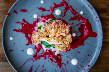 Roasted cumin cauliflower steak is elegantly presented with vibrant splashes of red beet puree and delicate white dots, creating a visually stunning culinary masterpiece.