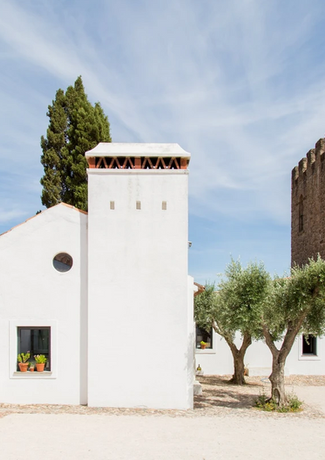 A white walled chimney, connected to a small house in the sandy courtyard with olive trees and flanked by an old stone tower with a round wooden door.