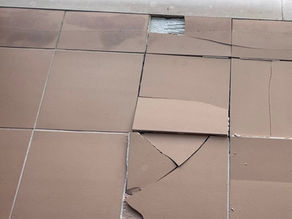 A high-angle photograph of a tiled balcony floor where several large, brown ceramic tiles have cracked and detached from the surface. The tiles are lifted and broken due to pressure, showing the concrete adhesive beneath where the tiles have failed.