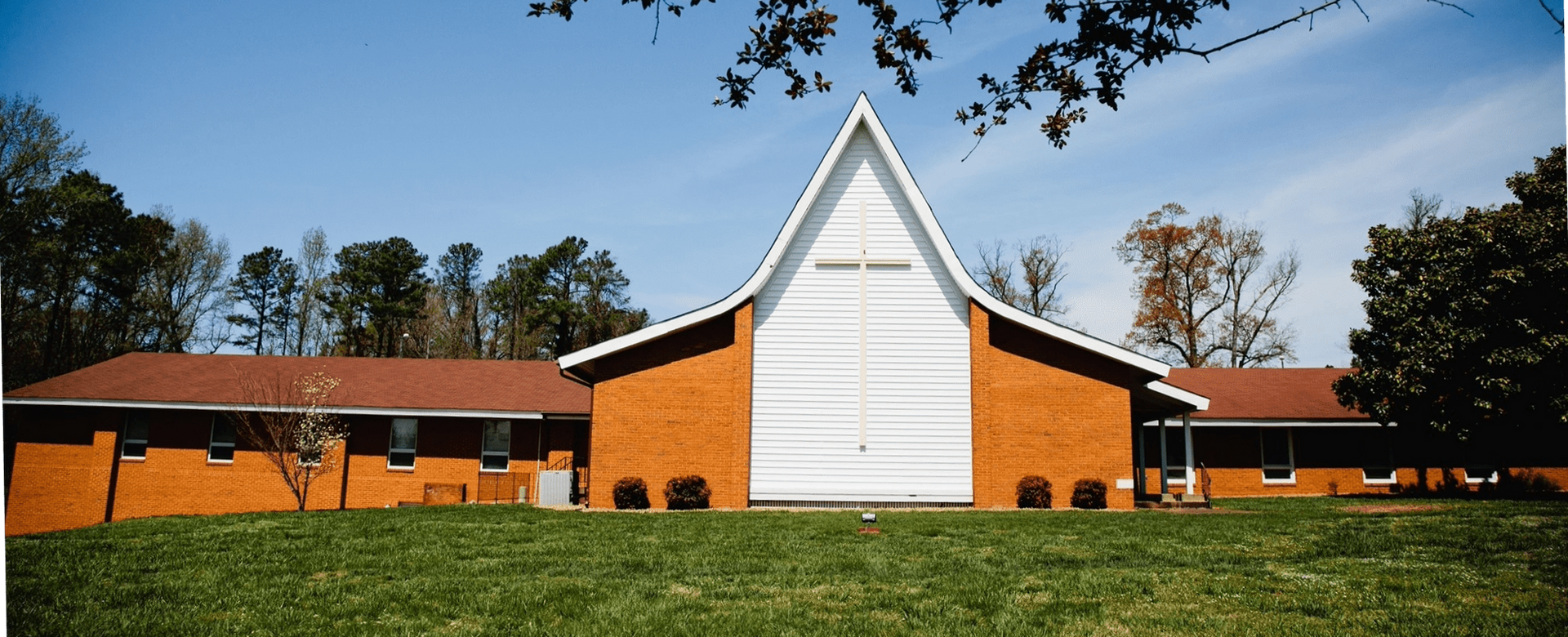 Brick church with tall white steeple, red roof wings, grassy yard, and oak trees on sides.