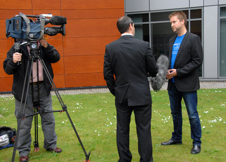 Our founder Pete stands on a grassy verge in front of a brick building being interviewed for TV