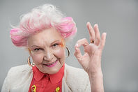An elderly lady with pink hair and glitter on her face looks at the camera and makes the 'ok' sign with her hand.