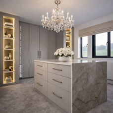 Luxury custom dressing room by KV Designs Leeds, featuring light grey bespoke fitted wardrobes, a central drawer island with a marble countertop waterfall edge, an elegant crystal chandelier, and integrated warm LED open shelving.