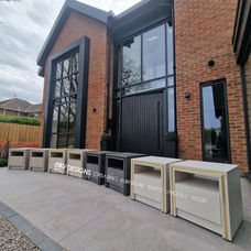 A row of luxury custom-made bedside cabinets by KV Designs Leeds lined up outside a modern luxury brick house with large black panoramic windows, highlighting a premium manufacturing run for high-end new build projects.