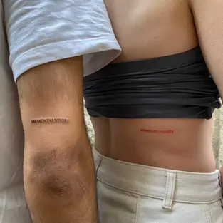 Two people showing matching fine line tattoos on their skin that read 'MEMENTO VIVERE' in elegant, minimalist typography. The person on the left has black ink on their inner arm, while the person on the right has red ink on their side/rib area. The photo is taken in a bright, natural setting in Budva, Montenegro