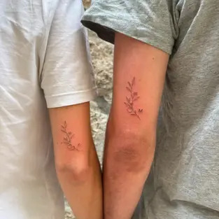 Fine line matching couple tattoo on the back of the arms featuring minimalist botanical leaves, a tiny flower, and personalized initials M and K.