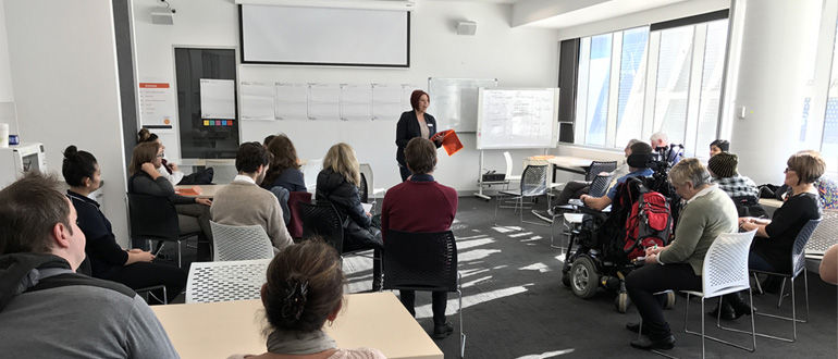 Wide landscape photo of a workshop room with a facilitator standing at the front speaking to a diverse group of seated adults, including a person using a wheelchair; neutral indoor colours with some red and dark accents; wide landscape aspect ratio.