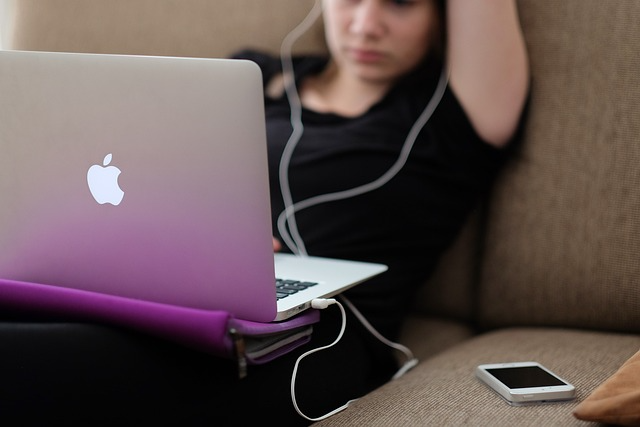 Woman seated on a sofa looking into an Apple laptop with an iPhone beside it and wired earbuds going to her ears.