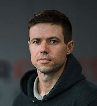 Close-up of a man with dark hair, wearing a black hoodie, standing against a blurred background