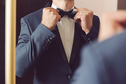 Man standing in front of a mirror in a tuxedo suit, fixing his bow tie.
