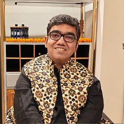 Nipun Malhotra with glasses smiling warmly, wearing a floral patterned vest over a black shirt. Background features marigold flowers and decorative items.