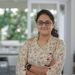 Bhumika Modh with glasses and a confident smile stands with folded arms, wearing a patterned top. Bright and airy background suggests a modern setting.
