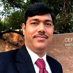 Subhash Chandra Vashishth in a suit and red tie stands outdoors, smiling confidently. Background features a lush green tree and a brick wall with partial text visible.