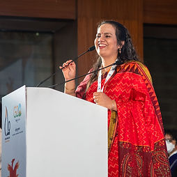 Nidhi Goyal in a red patterned sari smiles while speaking at a podium with two microphones, wearing a conference lanyard and standing in a wood-paneled indoor venue with a G20 logo visible on the lectern.