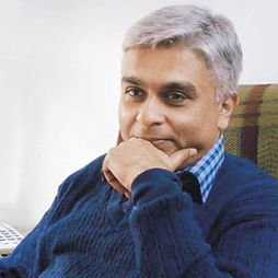 George Abraham with gray hair in a cozy blue sweater sits thoughtfully with chin resting on hand, conveying a calm and contemplative mood against a neutral background.