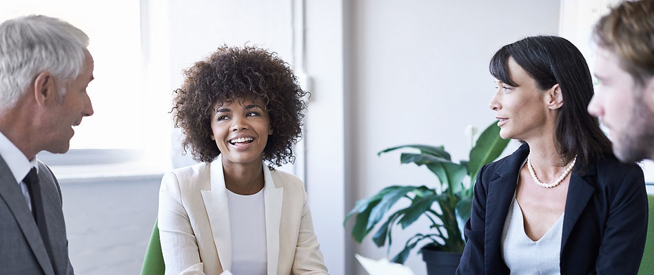 Female worker at the centre of the image smiles at a male collegue in an office setting. Two other colleagues smile back at her.