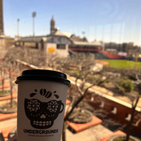 Hand holding "UNDERGROUND" coffee cup, sugar skull logo, blurred city view.