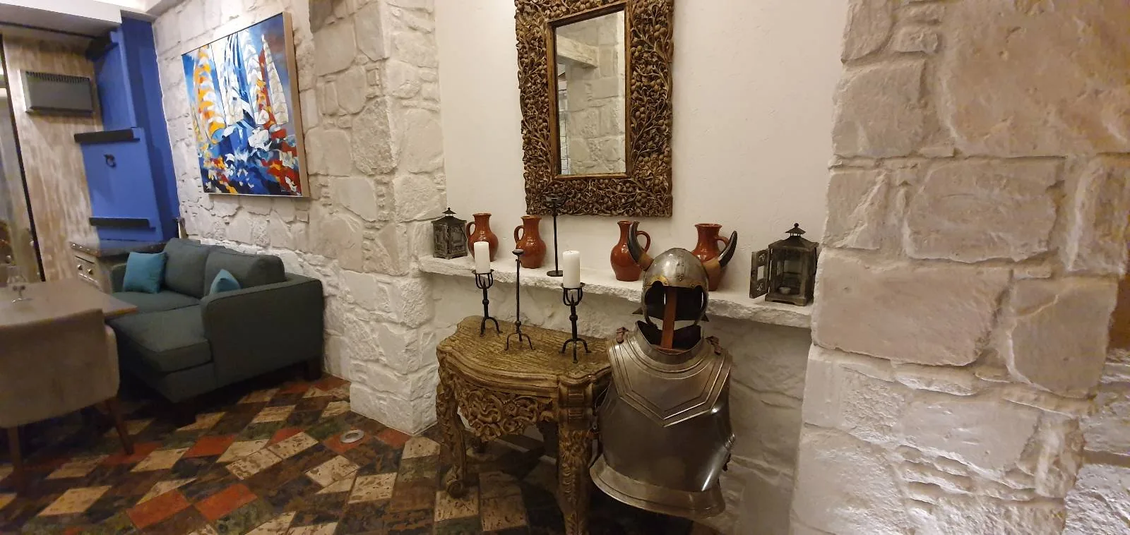 A room with white stone walls features a decorative suit of armor and a wooden table with candlesticks and vases. There's an ornate mirror above the table and a colorful abstract painting. A green couch and a chair are in the corner on a patterned floor.