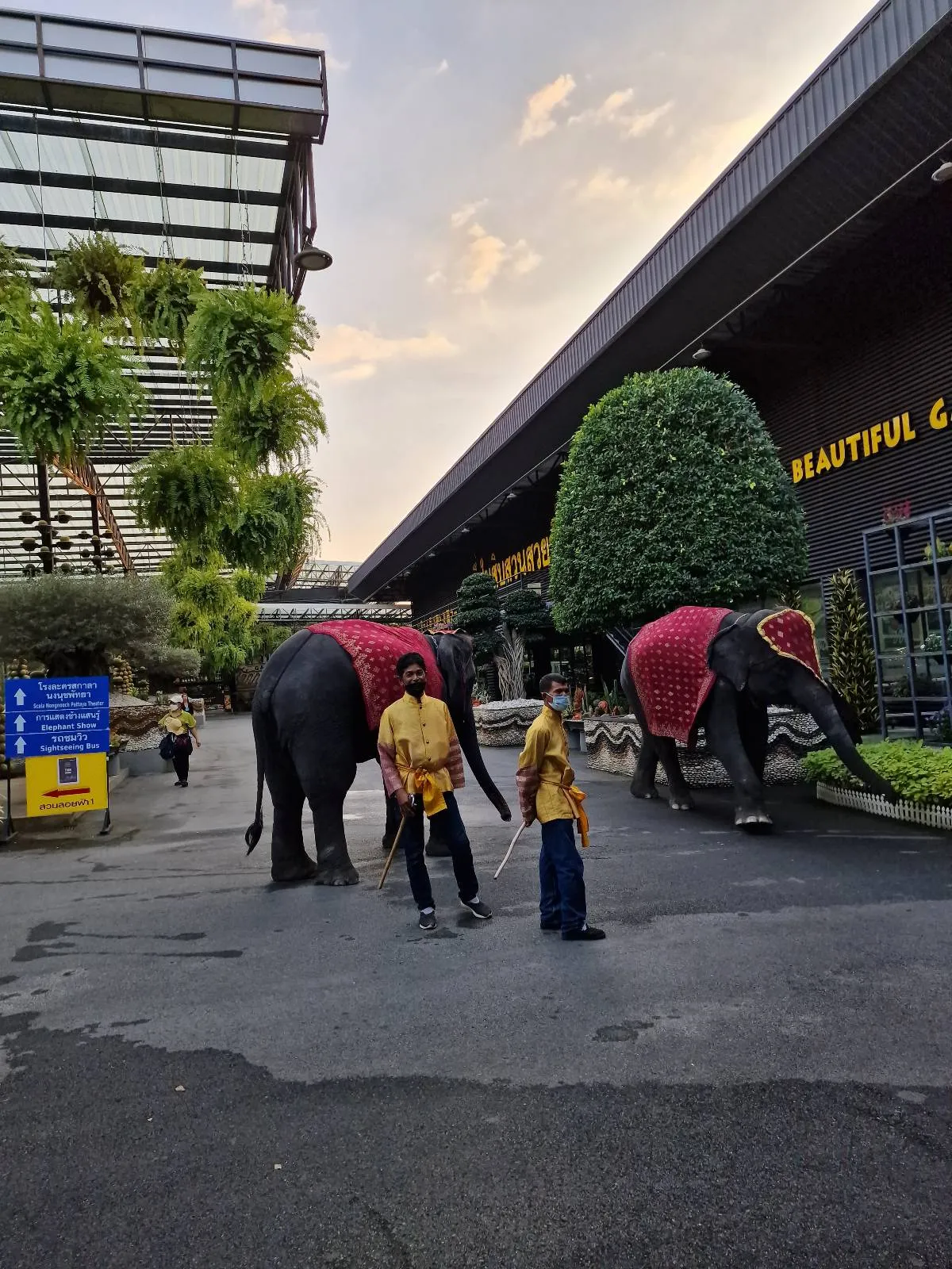 Two people standing in front of two elephants adorned with red cloths. The scene is set near a building with plants and a sign in the background. The sky is partly cloudy, and the area appears to be part of a tourist attraction.
