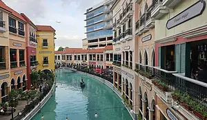 A scenic outdoor view of the Venice Grand Canal Mall in Manila, featuring a turquoise man-made canal with a gondola and gondolier, flanked by Mediterranean-style buildings in pastel yellow, pink, and red.