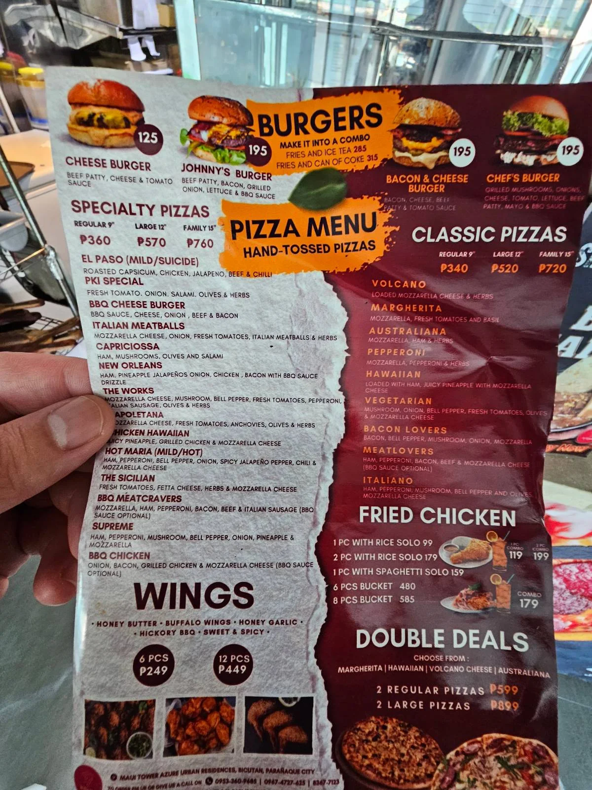 Close-up of a menu featuring burgers, specialty pizzas, classic pizzas, wings, fried chicken, and double deals. The menu has colorful images and descriptions of each item with prices. A hand is holding the menu.