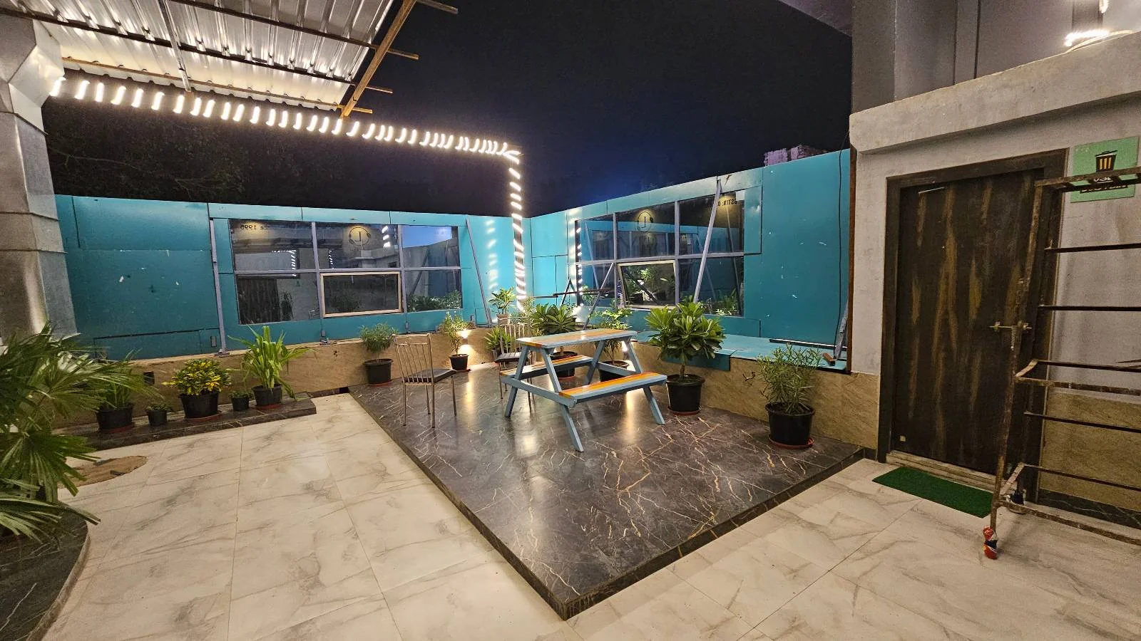 A nighttime rooftop patio features string lights, potted plants, and a seating area with a table and benches on a wooden platform. Blue glass panels line the edges, and a closed wooden door is on the right.