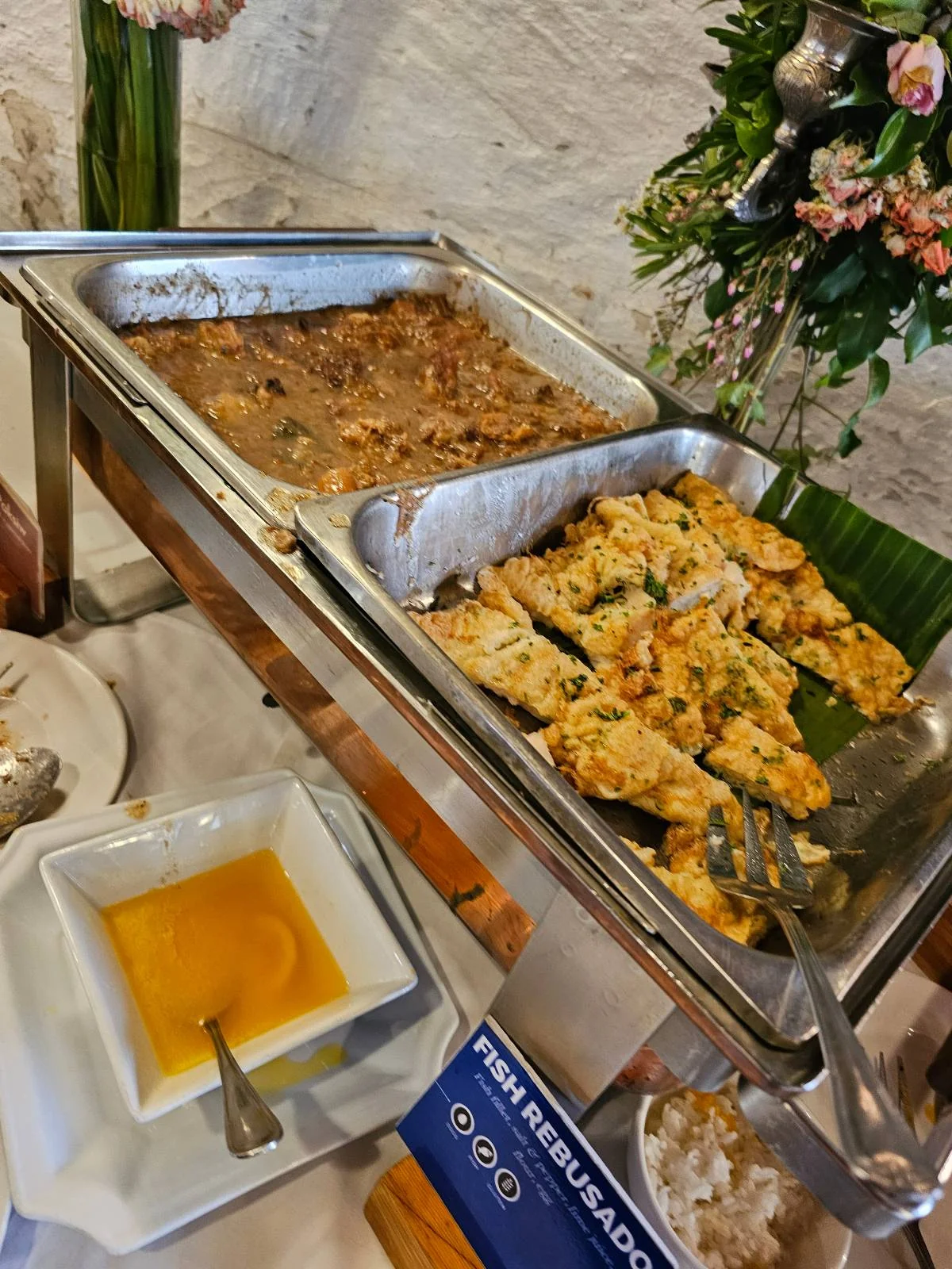 Buffet setup featuring trays of cooked meat and sauce, with a dish of yellow sauce on the side. A floral arrangement is partially visible in the background.