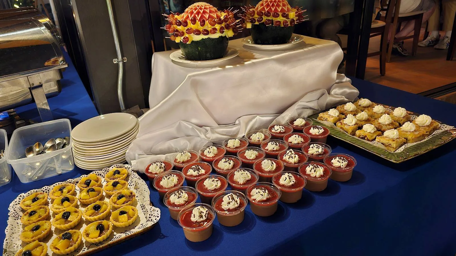 A dessert table with assorted treats, including cups with toppings, small cakes, and two fruit arrangements.