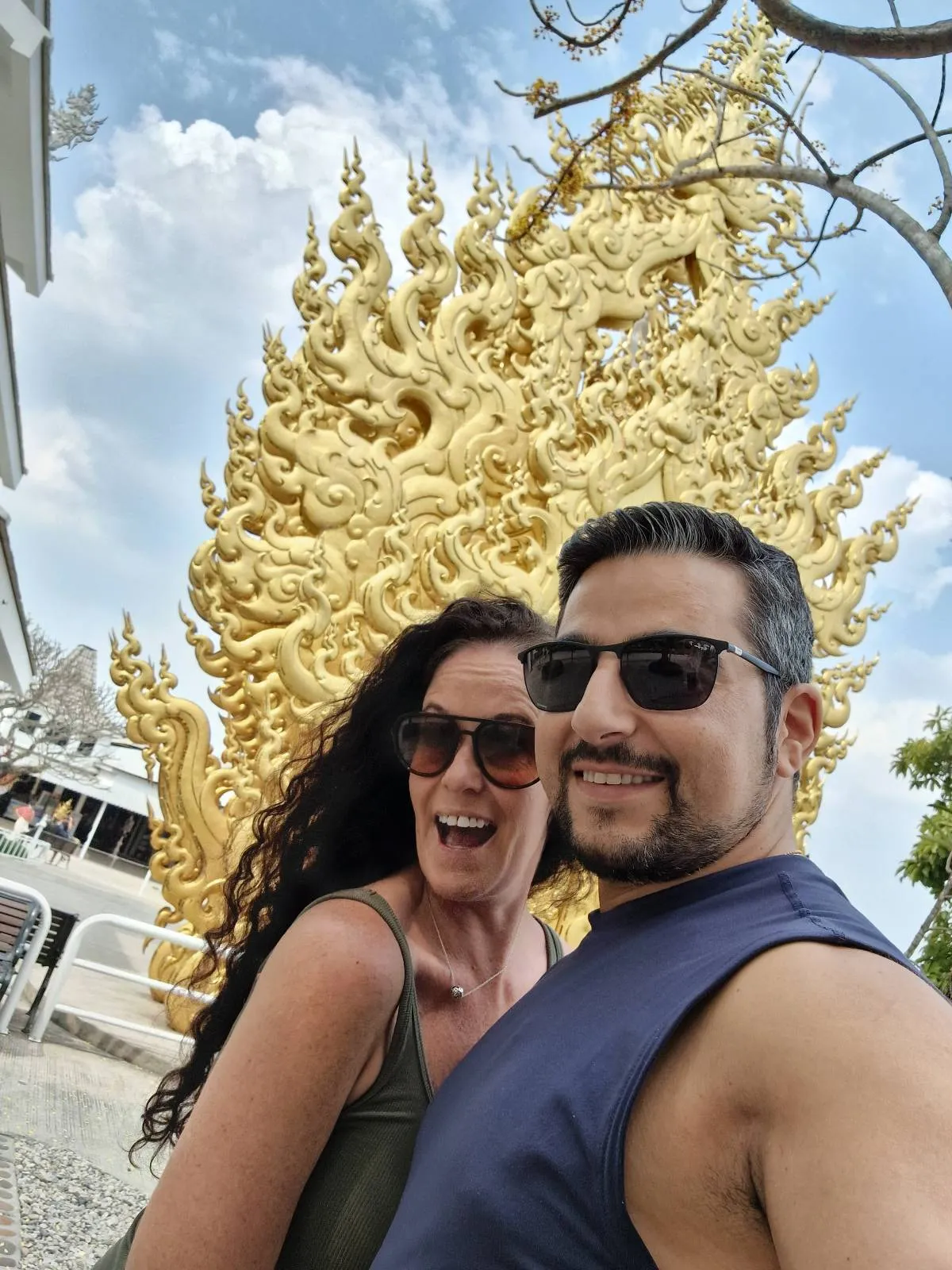 A man and woman wearing sunglasses are smiling in front of an ornate golden structure with intricate designs. The sky is partly cloudy, adding a bright backdrop to the scene.