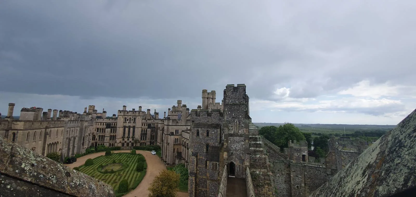 A sprawling stone castle with multiple turrets and towers under a cloudy sky. The foreground features a manicured garden and pathway, surrounded by the fortress walls. The landscape extends into a distant, lush horizon.